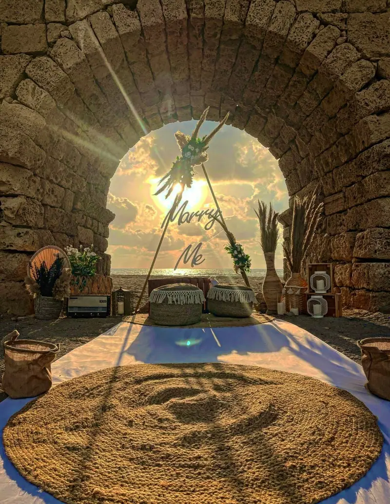 טיפי עם שלט Marry Me ושטיח קש בתוך קשת אבן עתיקה, כשהשמש שוקעת מאחור