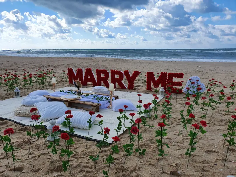 אותיות MARRY ME אדומות על החוף מאחורי שולחן פיקניק נמוך עם כריות לבנות, וורדים אדומים נעוצים בחול