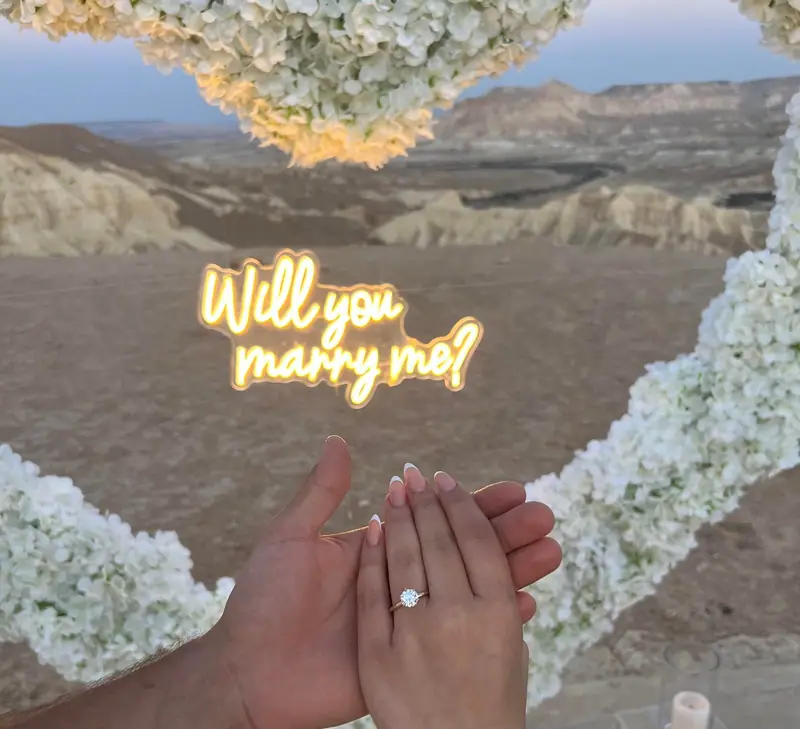 ידיים עם טבעת אירוסין מתחת לשלט ניאון Will You Marry Me