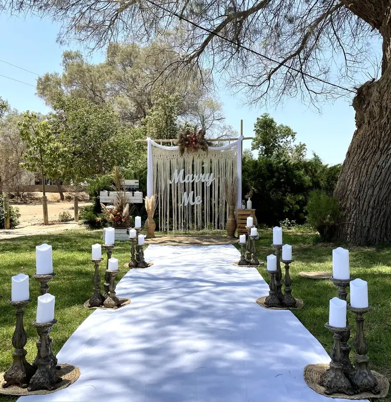 רקע מקרמה עם שלט Marry Me בקצה שביל לבן, עם נרות על מעמדי עץ בין עצי הפארק