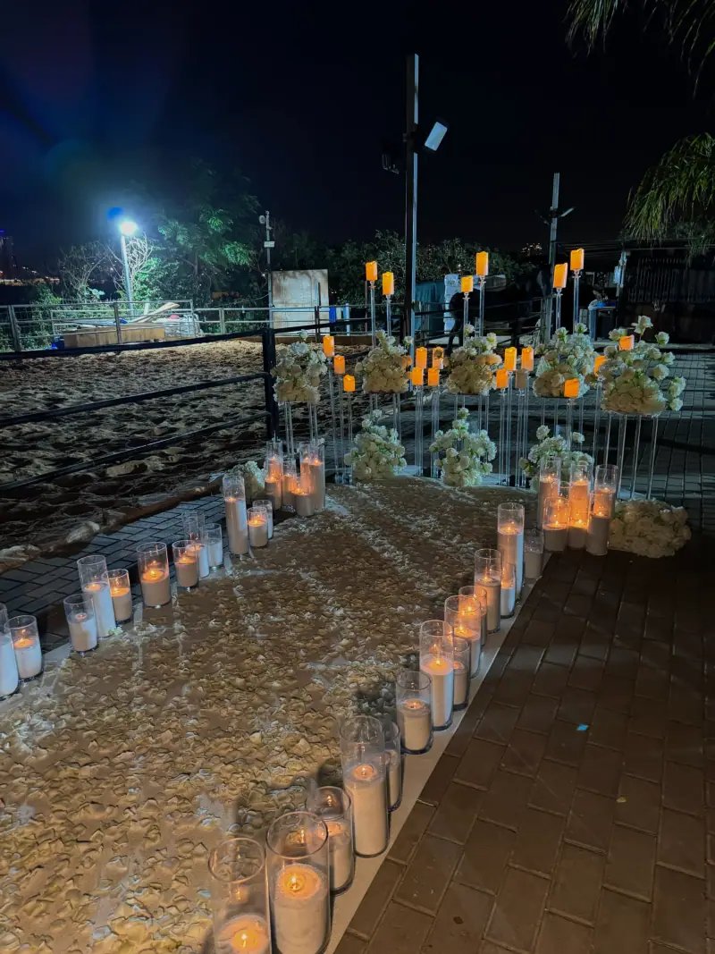 שביל לילי מוקף נרות בכוסות זכוכית וסידורי פרחים, על טיילת מול הים