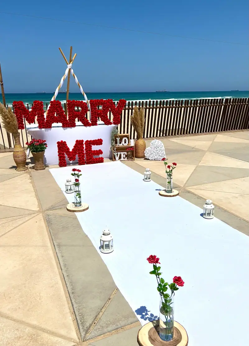 אותיות MARRY ME אדומות בקצה שטיח לבן עם ורדים אדומים, על גג מול הים