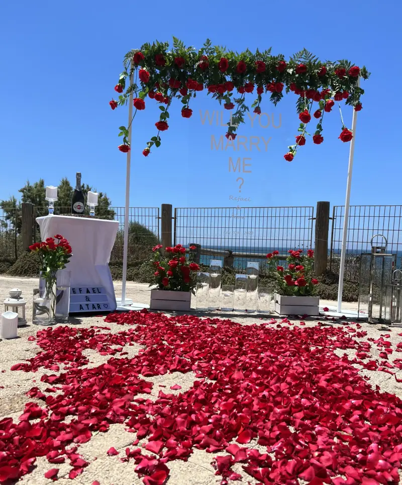 קשת לבנה עם ורדים אדומים בחלקה העליון ושטיח עלי ורדים אדומים, במרפסת מול הים
