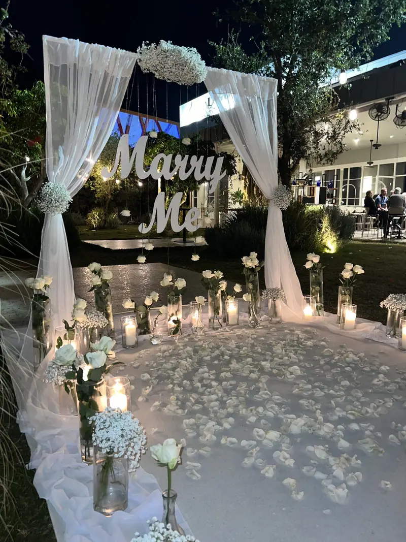 קשת עטופה בבד לבן עם אותיות Marry Me, נרות ועלי כותרת לבנים במרפסת בערב