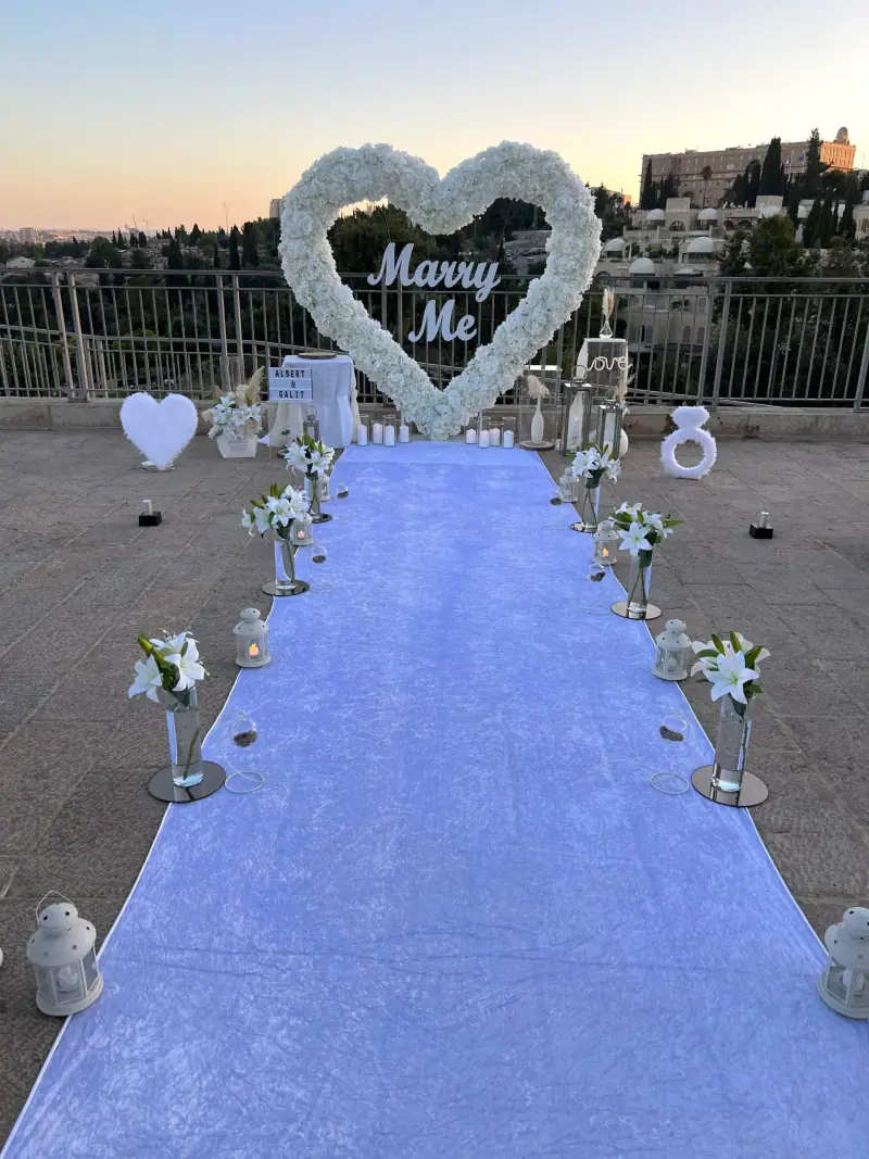 לב פרחים לבן עם כיתוב Marry Me בקצה שטיח לבן מוקף שושנים לבנות, על גג עם נוף לעיר