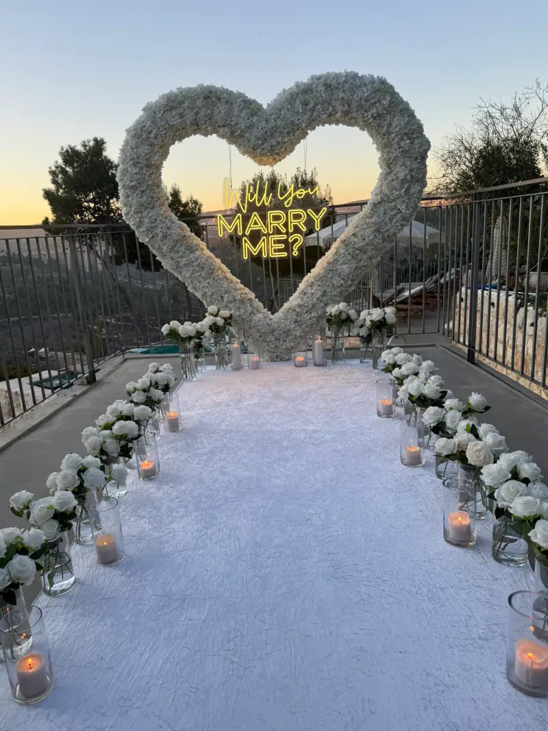 לב גדול מפרחים לבנים עם שלט ניאון Will You Marry Me, ושביל שטיח לבן מוקף פרחים ונרות על מרפסת בשקיעה