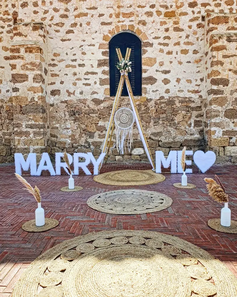 אותיות MARRY ME לבנות עם לב, וטיפי בוהו עם לוכד חלומות ושטיחים מול קיר אבן עתיק